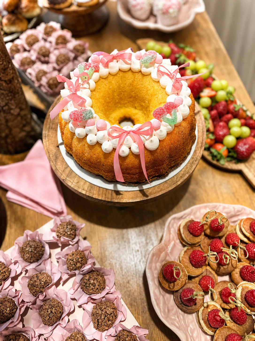 Bolo artesanal decorado em tons rosa, rodeado por fruta, brigadeiros e pequenas panquecas